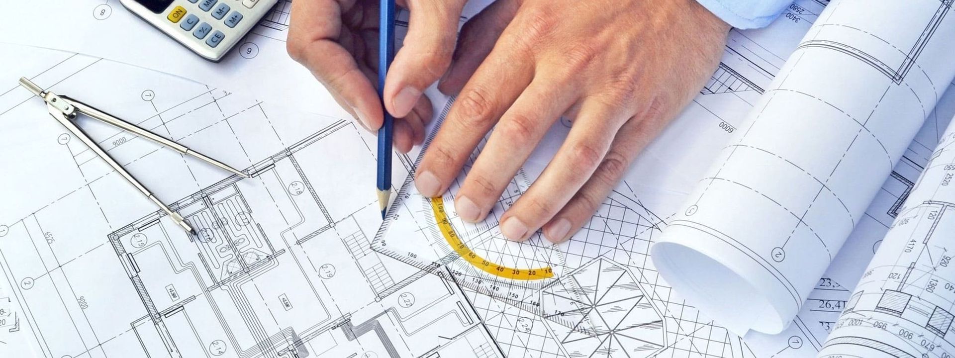 Eine Person hält einen Bleistift und ein Lineal über Baupläne, die auf einem Tisch ausgebreitet sind.